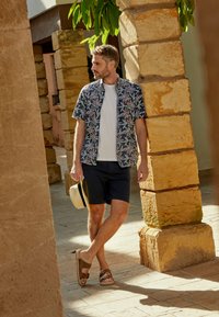 Een man draagt een donkere, bloemige overhemd met korte mouwen over een wit T-shirt, in combinatie met marineblauwe shorts en bruine sandalen, terwijl hij een strohoed vasthoudt.