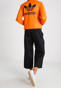 orange fleecejacka med svart Adidas-logga på ryggen, i kombination med svarta korta byxor och vita sneakers med ljusbruna sulor.