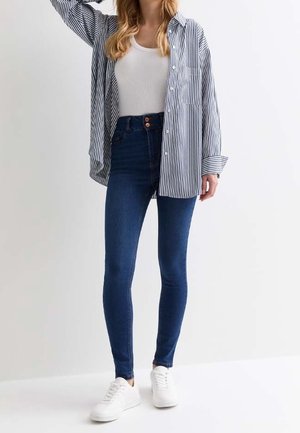 Chemise à rayures boutonnée, débardeur blanc, jean skinny bleu foncé, et baskets blanches. Coupe décontractée, style casual, poche avant sur la chemise.