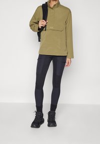 Olivgrön pullover med blixtlås och ficka fram, i kombination med svarta leggings-liknande byxor med texturerade paneler och en figursydd design.