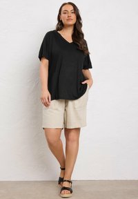 Donna in piedi contro un muro bianco, con una maglietta nera a V larga, pantaloni beige fino al ginocchio e sandali con zeppa neri, con la mano in tasca.
