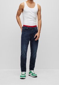 Top sin mangas blanco, vaqueros oscuros ajustados con una cinturilla roja de HUGO, y zapatillas verdes y blancas con cordones. Diseño casual y ajustado.