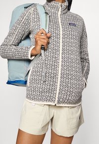 Osoba v mikině na zip s vzorem, béžových šortkách a se světle modrým batohy na rameni.