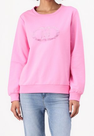 Kobieta ubrana w różową bluzę z długim rękawem z subtelnym wzorem na piersi, zestawioną z jasnoniebieskimi jeansami.