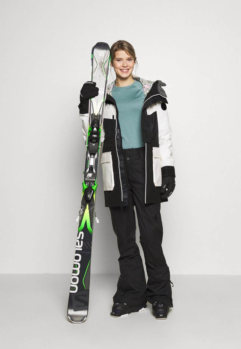 Skiausrüstung mit einer schwarz-weißen Jacke mit floralen Akzenten, schwarzen Skihosen und einem Paar Skier mit grünen Akzenten und Bindungen.