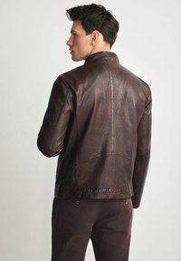 Mężczyzna zwrócony w lewo lekko się odwraca, ubrany w ciemnobrązową skórzaną kurtkę i ciemne spodnie, na tle jasnego, jednolitego tła.