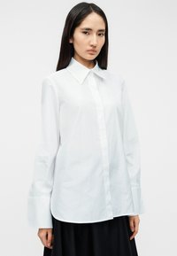 Λευκό πουκάμισο με μακριά μανίκια και μυτερό γιακά, κατασκευασμένο από λεία ύφανση. Διαθέτει ίσια γραμμή και φαρδιές μανσέτες, υιοθετώντας ένα μινιμαλιστικό σχέδιο.