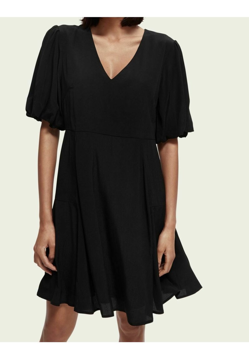 Femme portant une courte robe noire avec des manches bouffantes mi-longues et un décolleté en V, debout devant un fond clair uni.