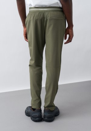 Mand iført olivengrønne slim-fit bukser med Calvin Klein-logo på taljebåndet og sorte sneakers, stående mod en ensfarvet lys baggrund.