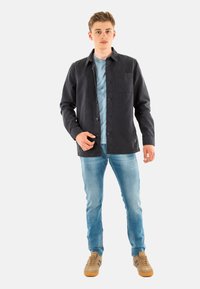 Donkerblauwe shirtjas met een borstzak, knoopsluiting en een gestructureerde afwerking, gedragen over een lichtblauw T-shirt en lichte jeans.