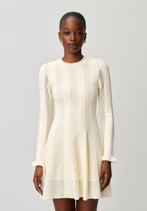 Mujer con un vestido de punto largo de color crema con rayas verticales y puños con volantes, de pie frente a un fondo gris claro liso.