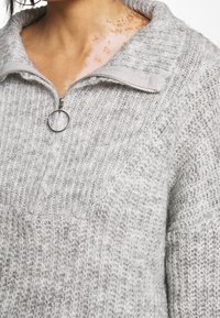 Maglione lavorato a maglia grigio chiaro con collo a zip quarti, tessuto testurizzato e un anello metallico per la zip. Presenta colletto e polsini a costine.