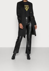 Schwarzer Mantel mit geradem Schnitt, Ein-Knopf-Verschluss und glatter Textur, kombiniert mit hoch taillierten schwarzen Kunstlederhosen und Ankle Boots.