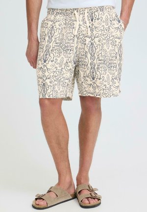 Beżowe szorty z granatowym wzorem kwiatowym i spiralnym, z wiązaniem w pasie, noszone z beżowymi sandałami na podwójnych paskach na stojącej osobie.