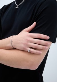 Hõbedane käevõru ristimustriga ja samasugune sõrmus väikeste kividega kaunistatud ristiga, kantud mustal T-särgil.