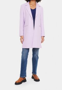 Cappotto su misura lilla con rever, tasche laterali, chiusura a un bottone, abbinato a jeans blu e stivaletti marroni.