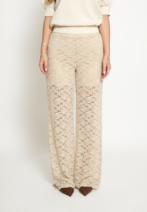 Woman wearing beige lace wide-leg pants, cream short-sleeve top, silver bracelets, and brown pointed-toe shoes standing against a plain background.