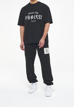 Persona che indossa una maglietta nera con la scritta "Trust the Process PEGADOR", pantaloni della tuta neri abbinati con toppa e scarpe da ginnastica bianche su sfondo bianco.