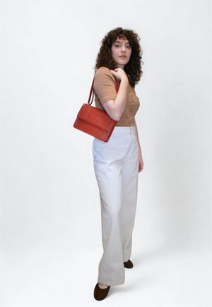 Chemise marron à boutons, pantalon blanc large, et petit sac rouge à motif crocodile. Chaussures marron foncé. Fond simple.