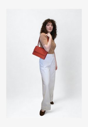 Chemise marron à boutons, pantalon blanc large, et petit sac rouge à motif crocodile. Chaussures marron foncé. Fond simple.