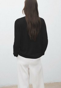 Black long-sleeve sweater with a round neckline, textured fabric, paired with white, tailored trousers, viewed from the back.