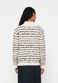 Femme aux cheveux bouclés foncés portant un pull crème avec des rayures horizontales noires texturées et une jupe plissée noire, vue de dos sur un fond uni.
