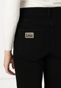 Mustat farkut, joissa on klassinen muotoilu ja takataskut. Huomionarvoinen harmaa "Lois" -labeli oikeassa taskussa. Sileä pinta.