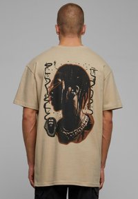 Beige cotton t-shirt featuring a large black graphic print of a profile, accompanied by abstract designs and a pharaoh's head illustration.