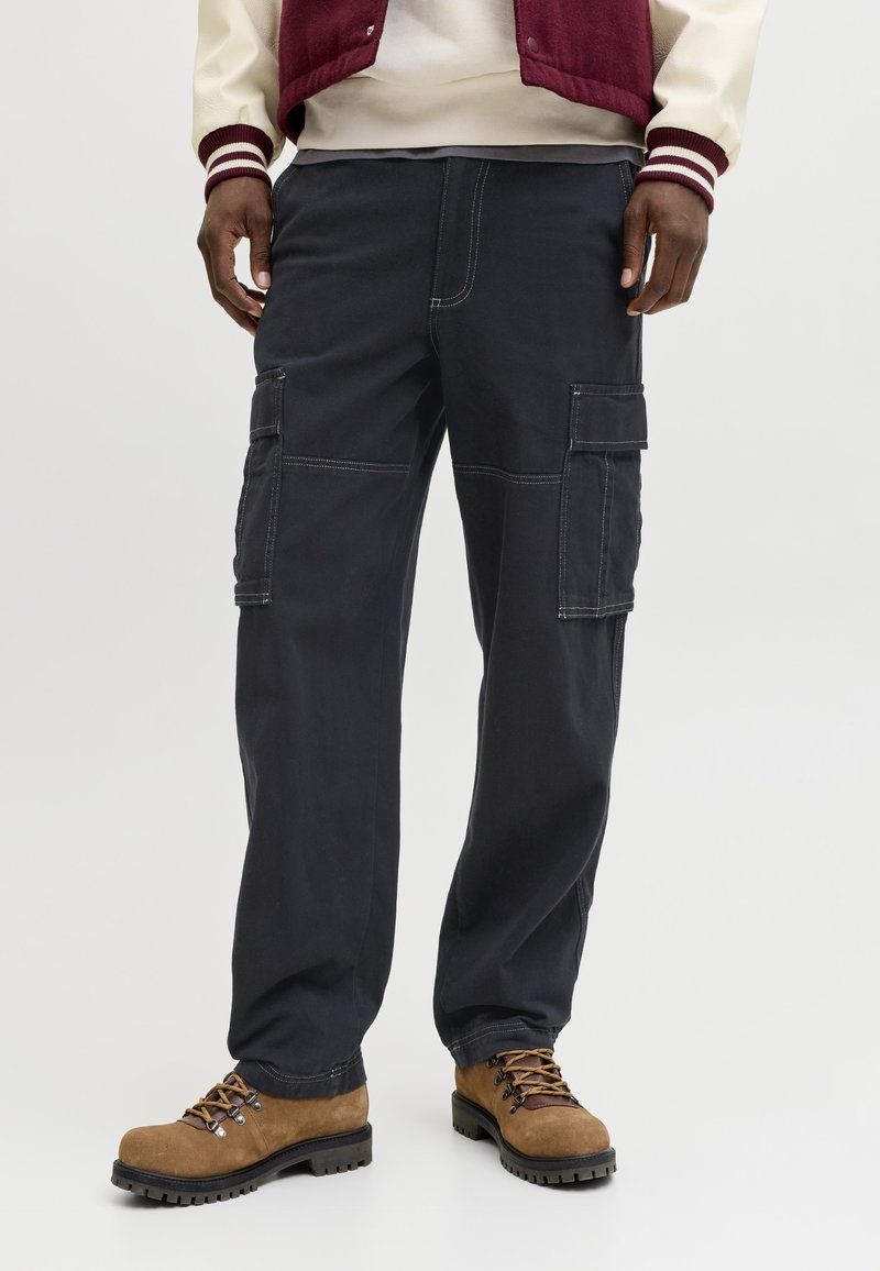 Homme portant un pantalon cargo noir avec plusieurs poches, des bottes de randonnée marron et une veste universitaire bordeaux et crème.