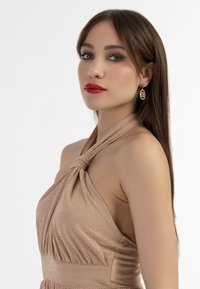 Bronzové šaty na halter s uzlovým detailem na výstřihu, texturovaná tkanina a volný, splývavý střih. Modelka má na sobě kruhové náušnice.