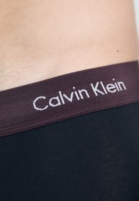 Čierne spodné prádlo so širokým vínovým pásom, na ktorom je bielym písmom napísané "Calvin Klein". Hladká textúra, priliehavý dizajn, minimalistický štýl.