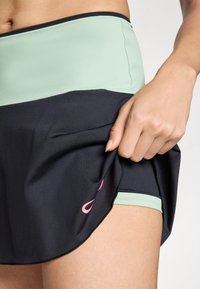 Falda atlética negra y menta con cintura alta, que presenta un logo rosa en el dobladillo. Fabricada con una tela elástica que absorbe la humedad.