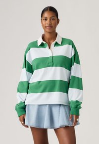 Γυναίκα που φοράει πράσινο και λευκό ριγέ μακρυμάνικο πουκάμισο ράγκμπι με λευκό γιακά και ανοιχτό μπλε πλισέ τζιν φούστα, στέκεται μπροστά σε ουδέτερο φόντο.