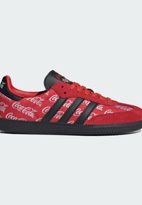Baskets rouges Adidas avec des bandes et une semelle noires, avec des logos blancs Coca-Cola en motif sur la tige en tissu et la zone en daim au niveau des orteils.