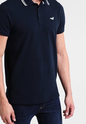 Granatowa koszulka polo z białym paskowanym kołnierzykiem i małym białym logo ptaka na lewej piersi, noszona z ciemnoniebieskimi dżinsami.