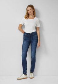 Obcisłe jeansy w ciemnoniebieskim denimu z subtelnym blaknięciem, zestawione z białym dopasowanym t-shirtem i jasnymi sneakersami. Prosty design i gładka faktura.