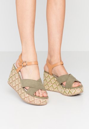 Wedge sandals featuring olive green suede cross straps, tan ankle straps, and a patterned woven heel in beige, green, and red accents.