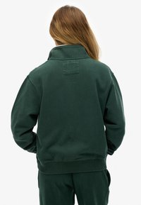 Felpa verde scuro con collo alto, tessuto strutturato e polsini e orlo a coste. Presenta una toppa quadrata sulla parte posteriore del collo.