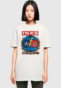 Vit oversized t-shirt med djärv grafiktryck som har blå och röda element, stjärnor, en jordglob, och texten "INXS CALLING ALL NATIONS."