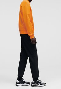 Oranssi huppari, jossa on ribbireunat, mustat kapenevat housut ja musta-valkoiset sneakersit, joissa on teksturoitu muotoilu ja nauhat, nähtynä sivulta.