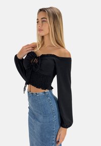 Schwarzes, schulterfreies Oberteil mit Raffung, langen Puffärmeln und verstellbarem Band. Kombiniert mit einem hochgeschnittenen Jeansrock.