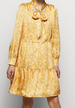 Robe jaune à manches longues avec petit imprimé floral, tissu plissé, col noué en nœud papillon et jupe à volants portée par une personne debout sur fond blanc.