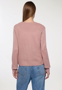 Różowy sweter z dzianiny z ściągaczem u dołu i przy mankietach, z okrągłym dekoltem i luźnym krojem. Noszony z jasnoniebieskimi dżinsami.