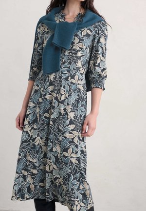 Kobieta w długiej sukience w kwiaty z rękawami 3/4 oraz niebieskim swetrze zarzuconym na ramiona na jednolitym tle.