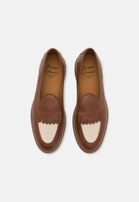 Mocasines de cuero marrón con un acento de flecos en forma de olas y detalles en panel crema. Costuras visibles a lo largo de la suela; el interior presenta el logo de la marca.