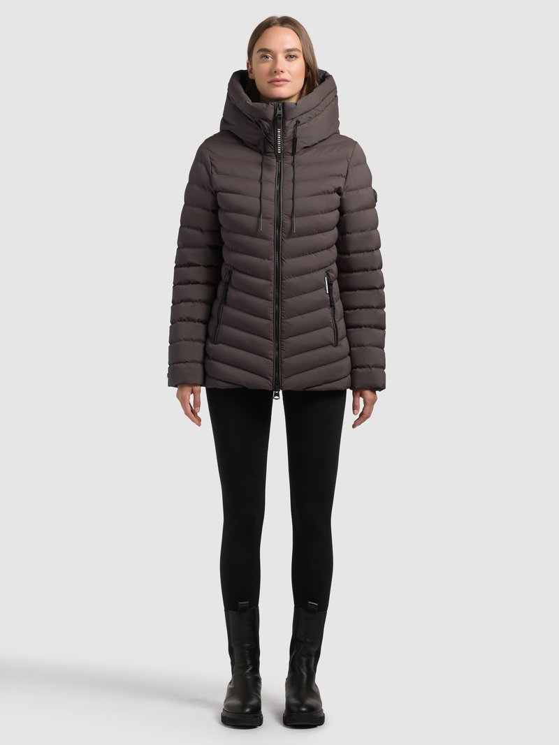 Giacca imbottita marrone scuro, con design a cuciture verticali, collo alto e chiusura frontale con zip. Indossata sopra leggings neri e stivali.