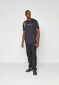 Musta urheilupaita, jossa on "TERREX"-logo, yhdistettynä mustiin housuihin ja tummiin lenkkareihin. Vihreä olkahihnareppu tuo kontrastia.