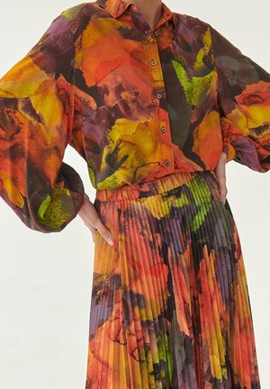 Person wearing a buttoned blouse and pleated skirt with an abstract watercolor pattern in orange, yellow, green, and purple tones.