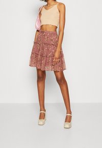 Femme portant un haut court beige, une jupe à volants mauve à fleurs, des escarpins crème Mary Jane et portant un petit sac à bandoulière rose.