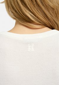 Blonde Person, der einen weißen Strickpullover trägt, von hinten gezeigt, mit einem kleinen gestickten Logo, das zentriert unter dem Rundhalsausschnitt angebracht ist.